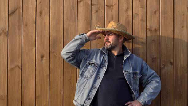 Man standing against wooden wall. Bearded. 40-45 years old. Dressed in denim jacket. Outdoors. Alone. Close-up. Portrait. Worker. Farmer. Builder.