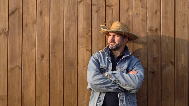 A man in a straw hat stands against a wooden wall. Dressed in a denim jacket. Bearded. Looking into the distance. Warm clothes. Farmer. Working man. Wooden boards in the background. Close-up. Serious look.