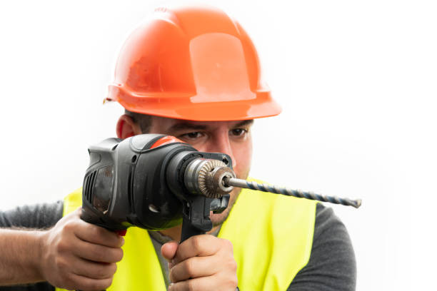 Close-up of adult male constructor wearing helmet and fluorescent vest using cordless power drill tool isolated on white background