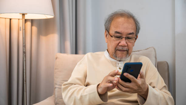 istock Asian old man engrossed reading a message in his mobile phone while sitting on a couch at house, Happy senior man sit in armchair relaxing with smartphone in living room at home 2185724610