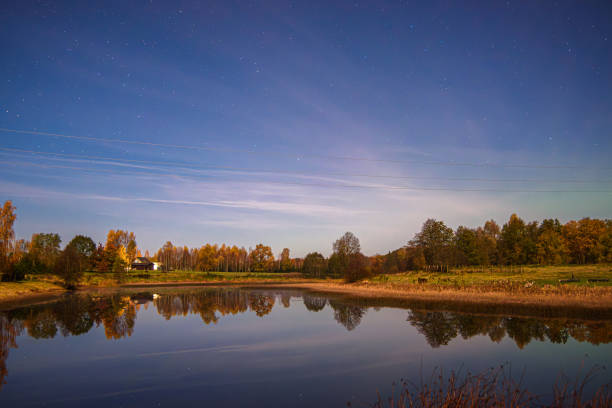 A serene rural pond under a starry sky at night, reflecting trees and a distant house surrounded by autumn colors. Peaceful atmosphere.