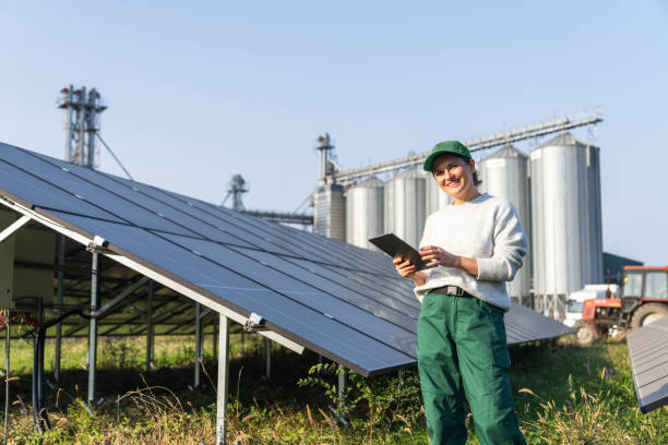 istock Female farmer wearing work uniform holds digital tablet in her hands stands next to solar panels. Modern agricultural farm using solar panels. Agricultural silos in the background. 2185737101