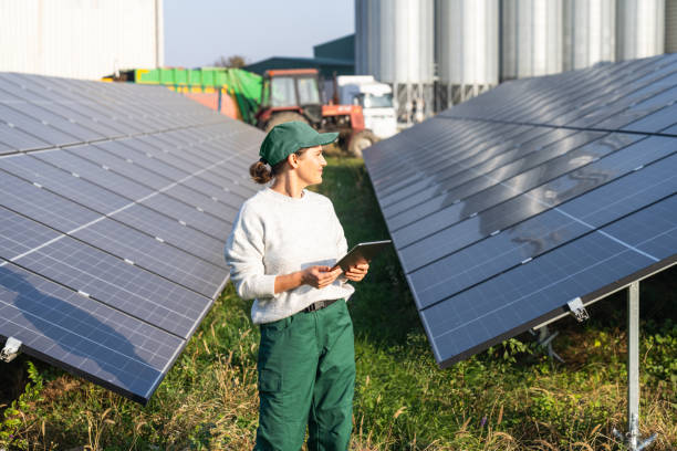istock Female farmer wearing work uniform holds digital tablet in her hands stands next to solar panels. Modern agricultural farm using solar panels. Agricultural silos in the background. 2185737504