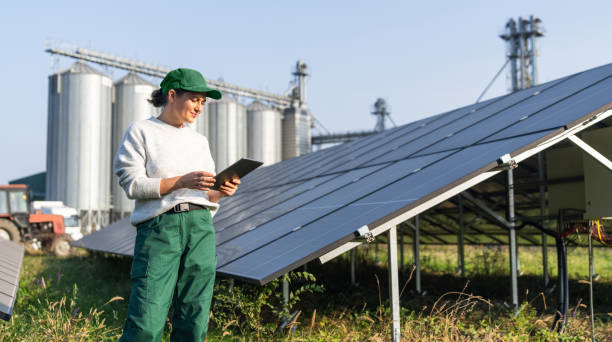 istock Female farmer wearing work uniform holds digital tablet in her hands stands next to solar panels. Modern agricultural farm using solar panels. Agricultural silos in the background. 2185739840