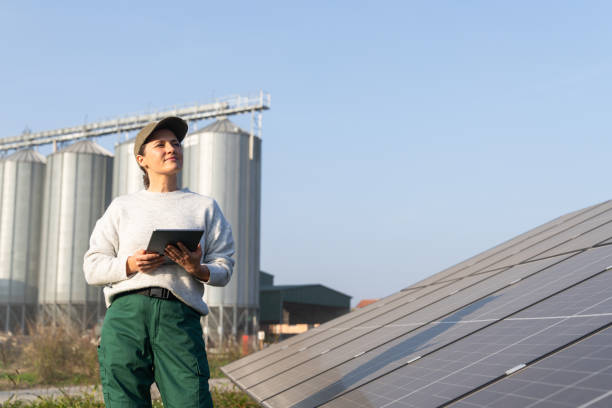istock Female farmer wearing work uniform holds digital tablet in her hands stands next to solar panels. Modern agricultural farm using solar panels. Agricultural silos in the background. 2185740641