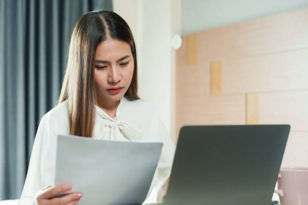 istock Asian woman holding paper document while working on laptop, white blouse, serious expression, seated in bedroom with modern wooden decor, warm lighting, illustrating work-from-home scenario. 2185742866