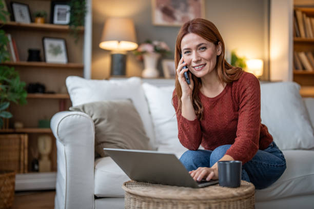 istock Cheerful woman using laptop and talking on mobile phone while sitting comfortably in her living room 2185746029