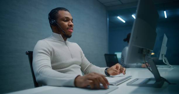 istock Call center office at night: African American technical support specialist in headset works on PC 2185752353