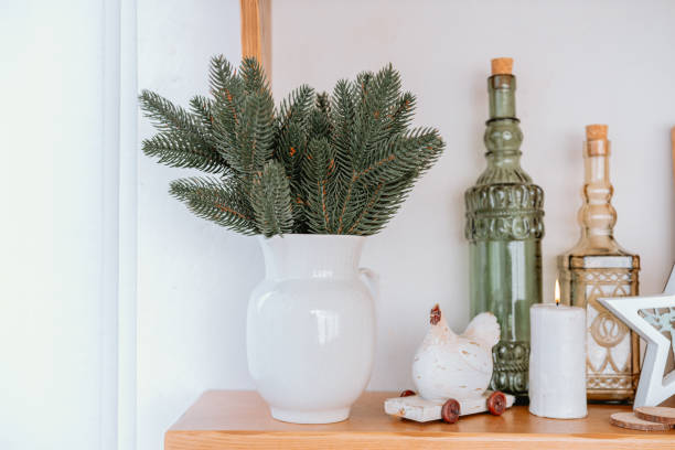 Decorative white vase with evergreen branches on wooden shelf at home