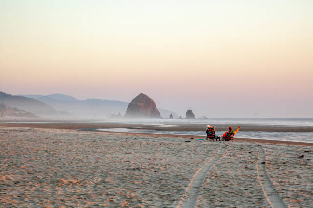 View across sandy beach to couple relaxing at sunset, island in ocean in distance, Haystack Rock, Oregon Islands National Wildlife Refuge
