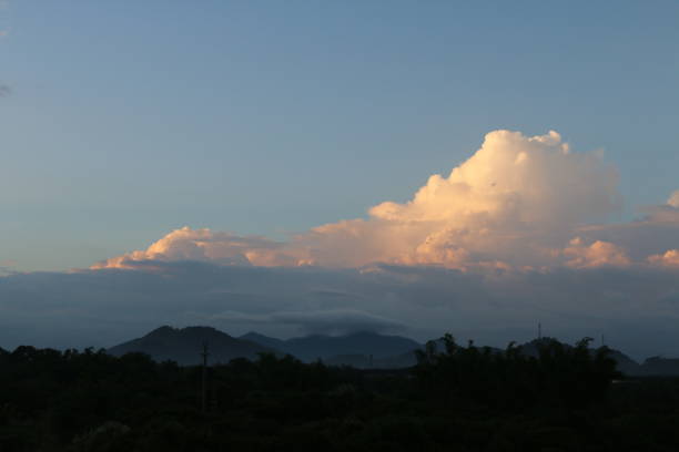 A stunning cumulonimbus cloud illuminated by the golden light of the setting sun. The cloud's warm hues contrast beautifully with the darker foreground, which features silhouettes of trees and houses in the village.