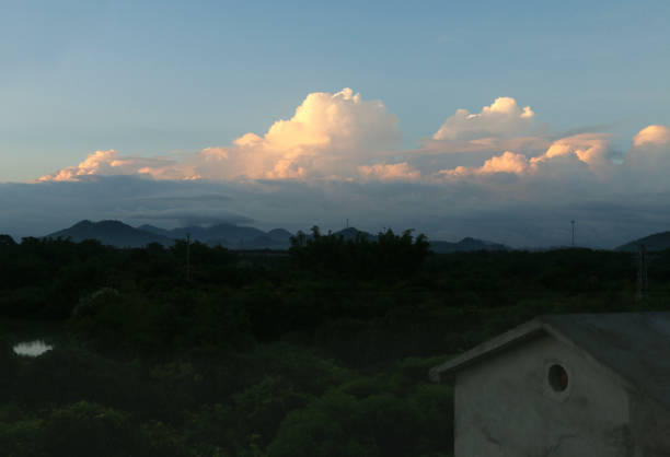 A stunning cumulonimbus cloud illuminated by the golden light of the setting sun. The cloud's warm hues contrast beautifully with the darker foreground, which features silhouettes of trees and houses in the village.