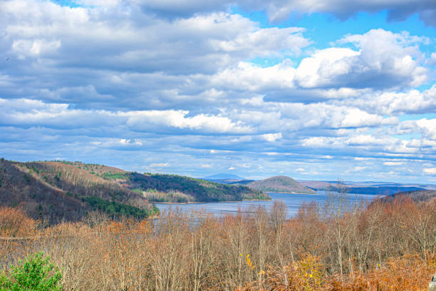 Serene autumn evening at a Pocono Mountains lake in Pennsylvania. Tranquil water mirrors dramatic cloudy sky with sunset hues. Shoreline features mix of evergreen and deciduous trees in fall foliage. Reeds and grasses emerge from shallow waters, adding foreground texture. Scene captures peaceful, contemplative essence of mountain autumn evening.