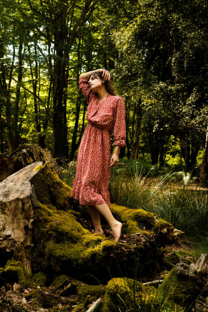 Fashion model posing barefoot on a mossy trunk in a green forest, touching her hair while wearing a red dress, enjoying the peace and quiet of nature