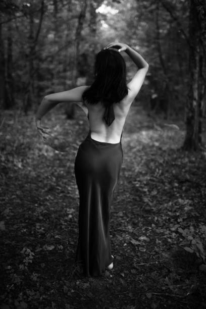 A woman in a dress stands amidst a tranquil forest. Mode standing back to camera and holding big round mirror in hand. Black and white image with selective focus. Image contains noise and motion blur