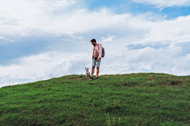 Man with backpack standing with small senior dog on green hilltop under cloudy blue sky