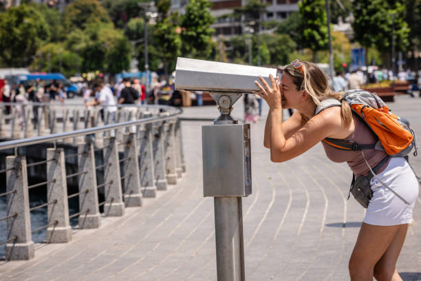 A woman leans over a coin-operated telescope, peering through to observe the surroundings on a sunny day, symbolizing curiosity and adventure in an urban setting.