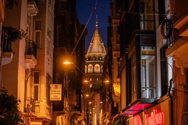 The iconic Galata Tower rises above a narrow street, framed by the evening's warm streetlights, with lively shops and signs adorning the city's vibrant nightlife.