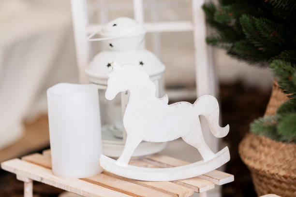 A white wooden rocking horse rests on a small wooden platform beside a candle and a lantern. The background features a festive tree, creating a warm and inviting atmosphere.