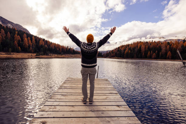 A man in a sweater and beanie stands on a wooden pier with arms outstretched, overlooking a peaceful autumn lake surrounded by colorful forests and mountains.