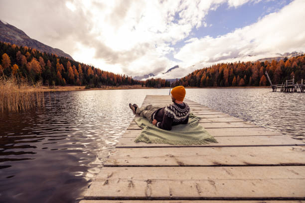 A man reclines on a wooden pier, wrapped in a green blanket, enjoying the serene autumn scenery with vibrant foliage and calm waters under a partly cloudy sky.