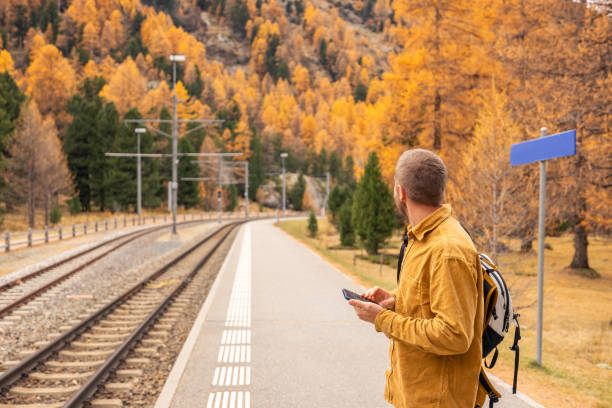 A man with a backpack smiles while using his smartphone at a railway station surrounded by vibrant fall foliage.
