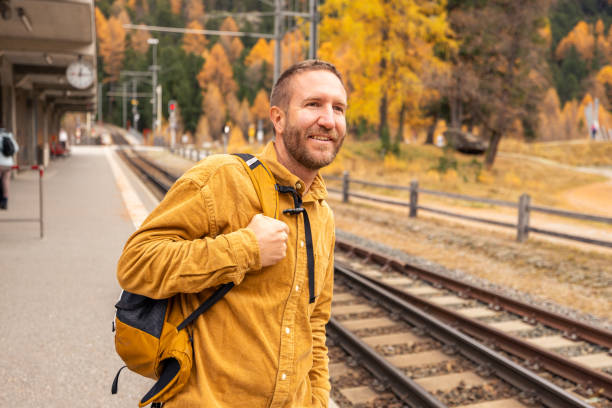 A cheerful man with a backpack stands on a train platform surrounded by stunning fall foliage, embodying the spirit of adventure and seasonal travel.