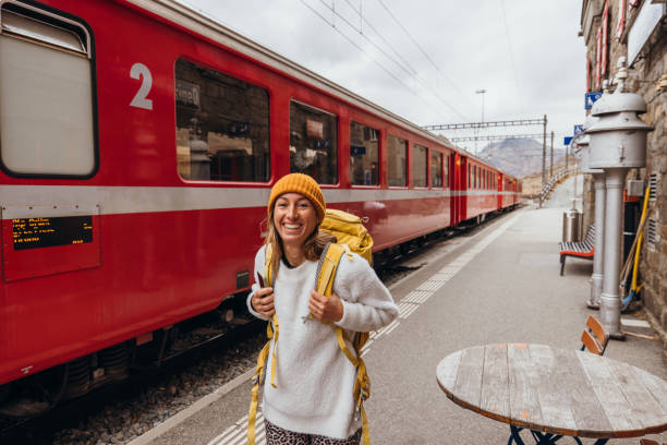 Woman with a yellow backpack standing on a platform beside a red train at a charming mountain station, ready for an adventurous journey.