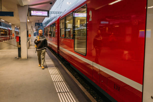 A cheerful man with a backpack stands at a train station next to a red train, ready for his journey. Evening lights enhance the station's modern atmosphere.