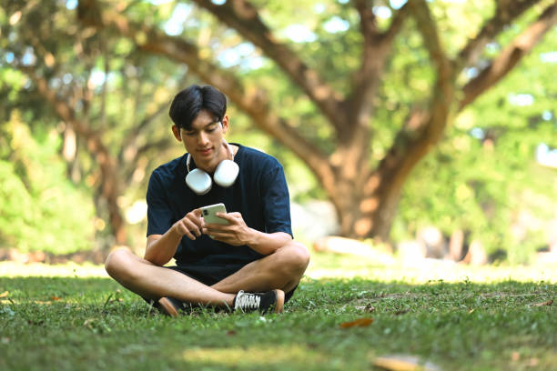 istock Young man sitting on the grass and using mobile phone in a park with sunlight filtering through the trees. 2185859429