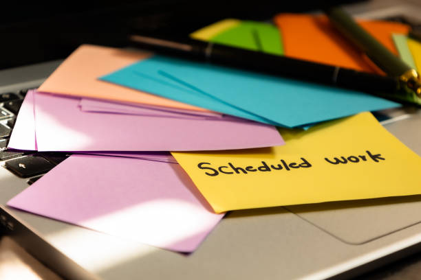 Close up of colorful sticky papers on laptop with pencils, pens on it and a yellow note that says scheduled work, work organization concept
