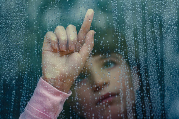Sad child looking through rainy window while pointing finger stock photo