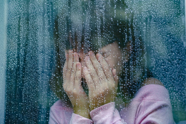 Sad child crying behind rainy window covering face with hands
