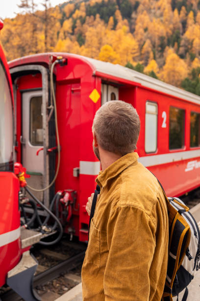 A traveler with a backpack in a mustard jacket stands beside a red train in an autumnal mountain landscape. Vibrant colors and serene surroundings enhance the scene.