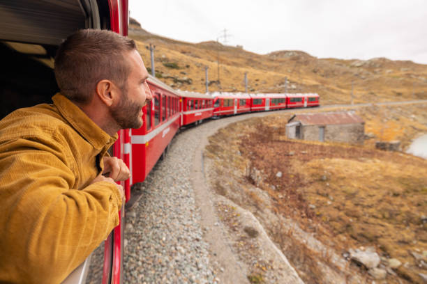 A man leans out of a red train window, soaking in the picturesque view of a winding railway alongside a serene lake and rugged mountains.