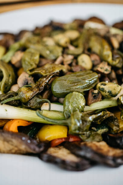 Close-up shot of colorful grilled vegetables featuring charred jalapeno peppers, sliced mushrooms, and fresh celery stalks arranged on a rustic wooden board. The vibrant dish showcases a mix of yellow and orange bell peppers in the background, with beautifully blistered green jalapenos and sautéed mushrooms in the foreground. The composition highlights the natural textures and colors of the vegetables, emphasizing their fresh, grilled appearance and appetizing char marks.