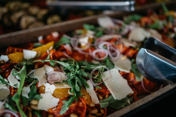 Close-up view of a rustic sheet pan filled with roasted butternut squash garnished with fresh arugula leaves, thinly sliced red onions, and shaved Parmesan cheese. The dish showcases vibrant orange squash pieces with caramelized edges, complemented by the peppery green arugula and purple onion rings. The white Parmesan shavings add texture and visual contrast to this healthy, seasonal dish photographed from above.