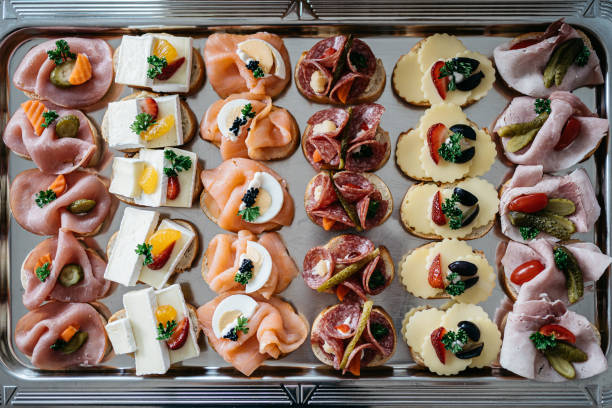 Overhead view of an elegant catering platter featuring assorted open-face sandwiches arranged in neat rows on a metal tray. The platter includes various toppings: smoked ham with pickles and carrots, brie cheese with citrus and strawberries, smoked salmon with caviar and cream, salami with cornichons, sliced cheese with berries and herbs, and turkey with cherry tomatoes. Each sandwich is artfully garnished with fresh parsley and decorative elements, making it perfect for events, parties, or upscale gatherings.