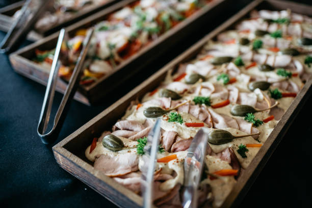 A sophisticated presentation of traditional Italian Vitello Tonnato featuring thinly sliced veal topped with creamy tuna sauce, garnished with fresh parsley, julienned carrots, and capers. The dish is artfully arranged in a long wooden serving board, showcasing the classic Italian appetizer with a modern twist. The delicate pink meat slices are beautifully draped and complemented by the pale, smooth sauce, creating an appealing contrast with the vibrant green herbs and bright orange carrots.