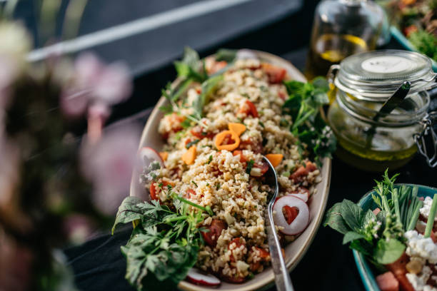 Close-up view of an elegant catering buffet display featuring a variety of fresh salads. The centerpiece is a beautifully presented grain salad with diced tomatoes, herbs, and mixed vegetables on an oval serving platter. Surrounding dishes showcase colorful mixed green salads with various toppings. The professional presentation includes serving utensils and small menu cards, suggesting a high-end catering or restaurant setting. The image is shot with selective focus, creating an appetizing depth of field effect.