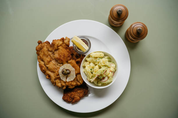 Traditional Austrian breaded veal cutlet served on a white plate with creamy potato salad as a side dish. The golden-brown, crispy schnitzel is garnished with a fresh lemon slice and capers. Wooden salt and pepper mills are visible in the background against a sage green surface, creating an authentic restaurant presentation of this classic European dish.