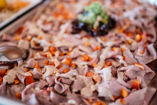 Close-up view of delicately sliced pink roast beef arranged on a serving platter with bright orange diced carrots scattered throughout. The meat is thinly sliced and layered, showing its tender, pink texture. In the background, there's a subtle hint of garnish with fresh herbs, creating an appetizing presentation perfect for catering events or buffet service. The image is shot with selective focus, emphasizing the detailed texture of the meat and the vibrant color contrast with the carrots.