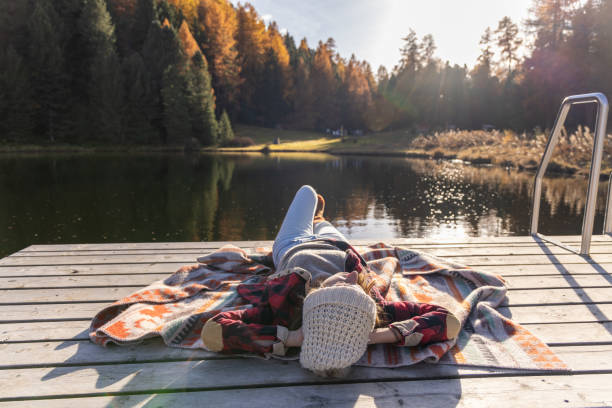 Woman in cozy attire lying on a blanket on a pier by a tranquil lake, soaking up the autumn sun with vibrant fall foliage and peaceful surroundings.