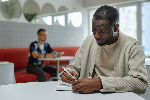 istock Black Man Planning Work Writing in Notebook while Sitting at Table in Office Center Cafe 2185912399