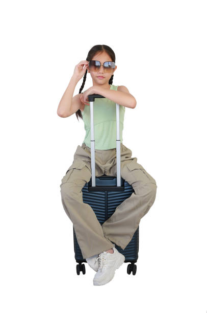 Asian girl child sitting on suitcase posing and looking straight camera while wearing sunglasses isolated on white background. kid fashion and travel concept. Full body photo.