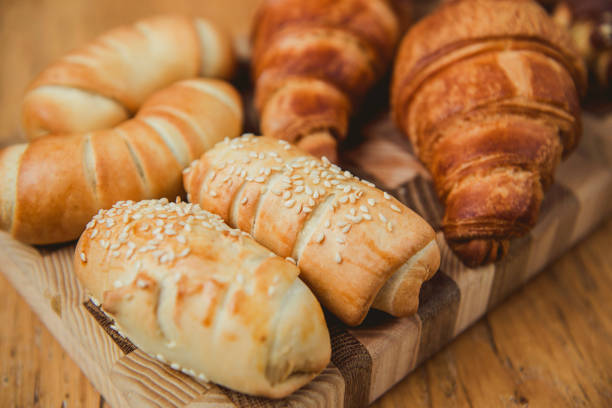 A close-up of a variety of freshly baked pastries, including golden croissants, sesame-topped rolls, and chocolate-dipped treats with crushed nuts. Placed on a rustic wooden cutting board, this image highlights a selection of sweet and savory bakery items.