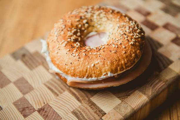 A close-up view of a sesame seed bagel filled with creamy cheese and slices of deli meat, placed on a rustic wooden cutting board. This image showcases a classic breakfast or lunch option, perfect for food photography or casual dining concepts.