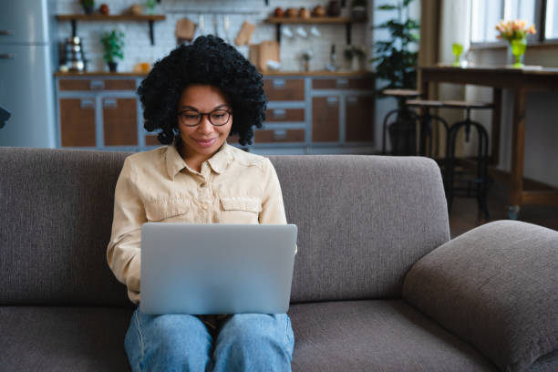 istock Smiling young black woman freelancer working from home, sitting on couch, using laptop, copy space. Relaxed african american lady enjoying her weekend, surfing on internet on laptop 2185928138
