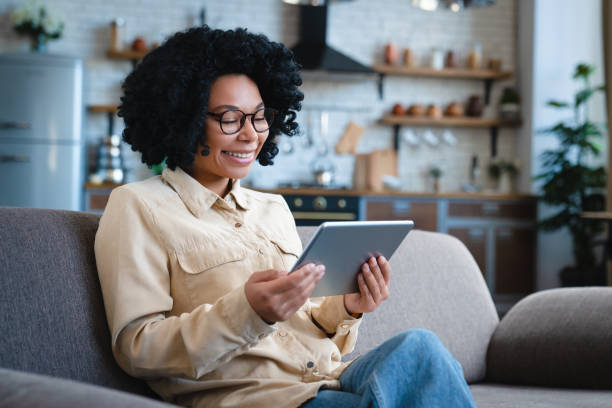 istock Happy young Black woman relaxing on sofa, using tablet computer, reading book, watching movie, media content, chatting online, talking on video call, laughing, smiling. Internet communication 2185928204