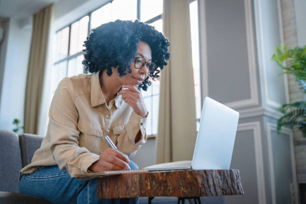 istock Young concentrate black woman using laptop at home and taking notes, happy african american female working remotely with computer while sitting on couch in living room at home 2185928253